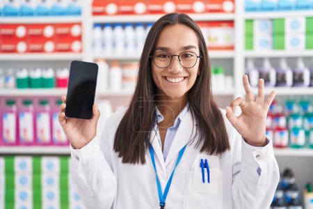 Téléchargez les photos : Jeune femme hispanique travaillant à la pharmacie pharmacie montrant écran smartphone faire signe ok avec les doigts, sourire geste amical excellent symbole - en image libre de droit