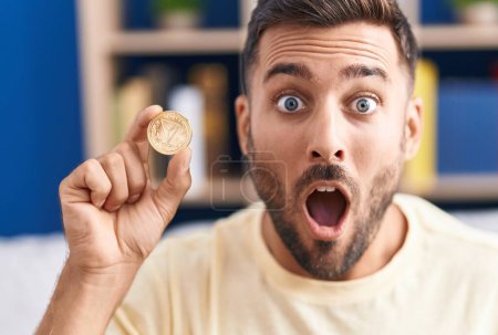 Foto de Hombre hispano guapo sosteniendo moneda criptomoneda tron asustado y sorprendido con la boca abierta para sorpresa, cara de incredulidad - Imagen libre de derechos
