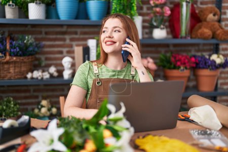 Téléchargez les photos : Jeune rousse femme fleuriste parler sur smartphone en utilisant un ordinateur portable à la boutique de fleurs - en image libre de droit