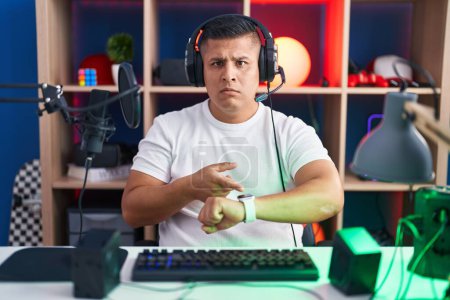 Téléchargez les photos : Jeune homme hispanique jouant à des jeux vidéo à la hâte pointant vers regarder le temps, l'impatience, bouleversé et en colère pour retard de délai - en image libre de droit