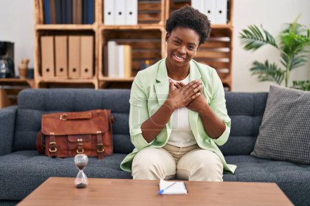 Foto de African american woman psychologist with hands on heart sitting on sofa at psychology center - Imagen libre de derechos