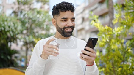 Téléchargez les photos : Jeune homme hispanique tenant une bouteille d'eau en utilisant un smartphone au parc - en image libre de droit