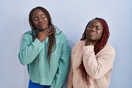 Foto de Dos mujeres africanas de pie sobre fondo azul tocando el cuello doloroso, dolor de garganta por gripe, clod e infección - Imagen libre de derechos