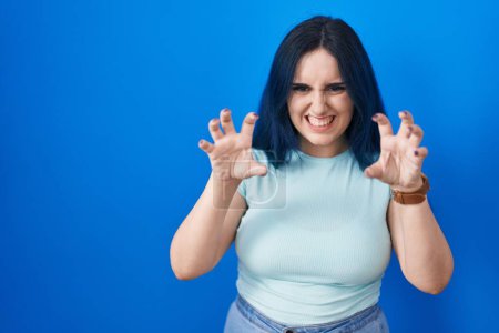 Photo for Young modern girl with blue hair standing over blue background smiling funny doing claw gesture as cat, aggressive and sexy expression - Royalty Free Image