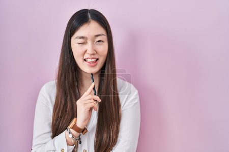 Photo for Chinese young woman holding pencil over pink background winking looking at the camera with sexy expression, cheerful and happy face. - Royalty Free Image