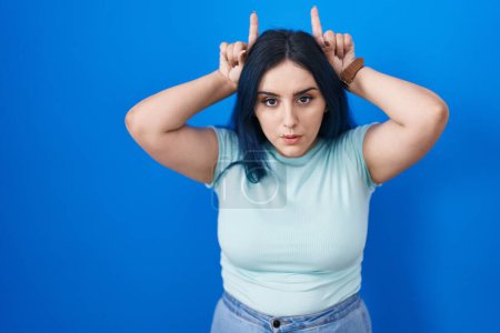 Téléchargez les photos : Jeune fille moderne avec les cheveux bleus debout sur fond bleu faisant geste drôle avec doigt sur la tête comme cornes de taureau - en image libre de droit