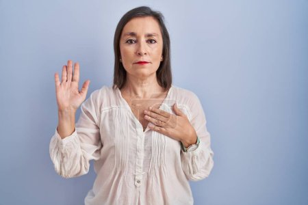 Téléchargez les photos : Femme hispanique du Moyen Âge debout sur fond bleu jurant avec la main sur la poitrine et la paume ouverte, faisant un serment de fidélité - en image libre de droit
