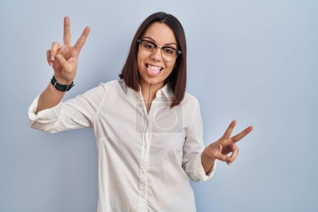 Téléchargez les photos : Jeune femme hispanique debout sur fond blanc souriant avec la langue montrant les doigts des deux mains faisant signe de victoire. numéro deux. - en image libre de droit