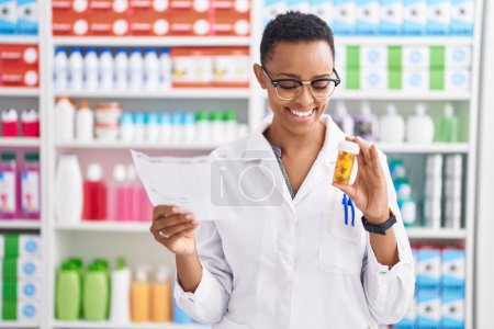 Foto de Farmacéutica afroamericana sosteniendo pastillas botella lectura prescripción en farmacia - Imagen libre de derechos