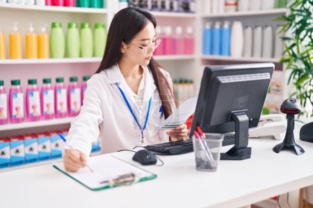 Photo for Young chinese woman pharmacist writing on document reading medical report at pharmacy - Royalty Free Image