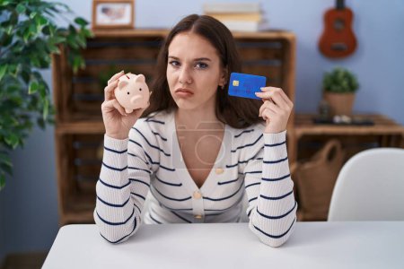 Foto de Chica hispana joven sosteniendo alcancía y tarjeta de crédito escéptico y nervioso, frunciendo el ceño molesto debido a un problema. persona negativa. - Imagen libre de derechos