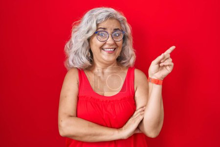 Téléchargez les photos : Femme d'âge moyen avec les cheveux gris debout sur fond rouge avec un grand sourire sur le visage, pointant avec le doigt de la main vers le côté regardant la caméra. - en image libre de droit
