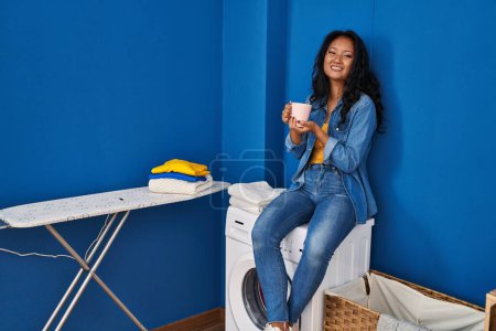 Téléchargez les photos : Jeune femme chinoise buvant du café en attendant la machine à laver à la buanderie - en image libre de droit