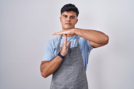 Foto de Joven hispano vistiendo delantal sobre fondo blanco haciendo un gesto de tiempo con las manos, rostro frustrado y serio - Imagen libre de derechos