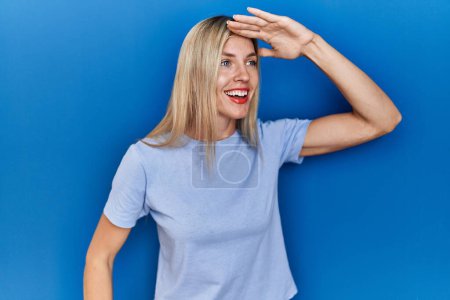 Foto de Hermosa mujer rubia con camiseta casual sobre fondo azul muy feliz y sonriente mirando lejos con la mano sobre la cabeza. concepto de búsqueda. - Imagen libre de derechos