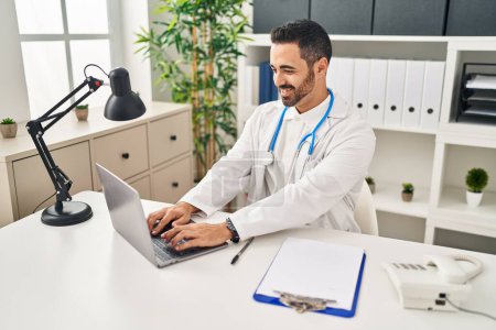Photo for Young hispanic man wearing doctor uniform using laptop working at clinic - Royalty Free Image