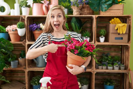 Foto de Joven mujer caucásica trabajando en floristería sosteniendo planta sonriendo feliz señalando con la mano y el dedo - Imagen libre de derechos