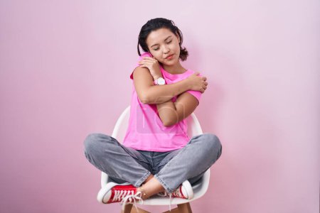 Photo for Hispanic young woman sitting on chair over pink background hugging oneself happy and positive, smiling confident. self love and self care - Royalty Free Image