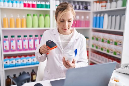 Foto de Joven mujer rubia farmacéutica escanear pastillas utilizando el ordenador portátil en la farmacia - Imagen libre de derechos
