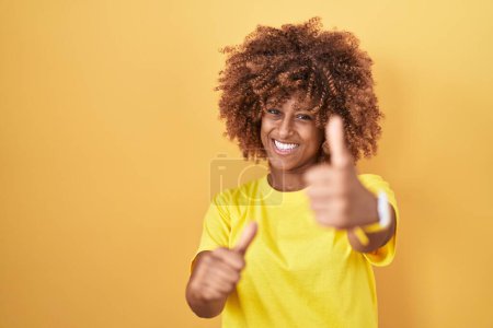 Foto de Mujer hispana joven con el pelo rizado de pie sobre fondo amarillo aprobando hacer gesto positivo con la mano, pulgares hacia arriba sonriendo y feliz por el éxito. gesto ganador. - Imagen libre de derechos