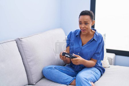 Foto de Mujer afroamericana jugando videojuegos sentada en el sofá en casa - Imagen libre de derechos