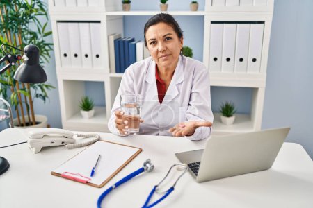 Photo for Hispanic mature doctor woman holding pill and glass of water relaxed with serious expression on face. simple and natural looking at the camera. - Royalty Free Image