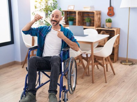 Foto de Hombre hispano con barba sentado en silla de ruedas sosteniendo nuevas llaves de la casa sonriendo feliz señalando con la mano y el dedo - Imagen libre de derechos