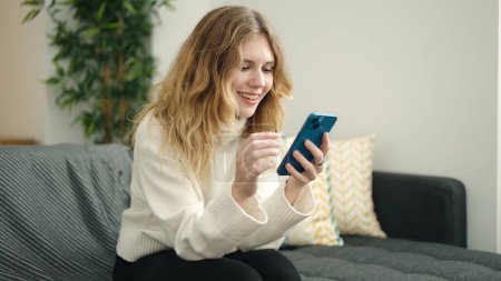 Foto de Mujer rubia joven usando teléfono inteligente sentado en el sofá en casa - Imagen libre de derechos