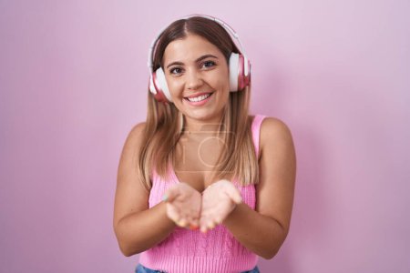 Foto de Mujer rubia joven escuchando música usando auriculares sonriendo con las palmas de las manos juntas recibiendo o dando gesto. retención y protección - Imagen libre de derechos