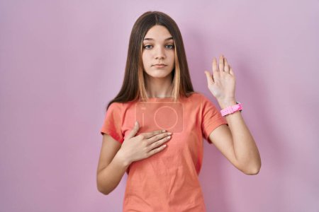 Téléchargez les photos : Adolescente debout sur fond rose jurant avec la main sur la poitrine et la paume ouverte, faisant un serment de fidélité - en image libre de droit