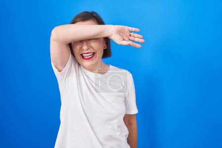 Foto de Middle age hispanic woman standing over blue background covering eyes with arm smiling cheerful and funny. blind concept. - Imagen libre de derechos