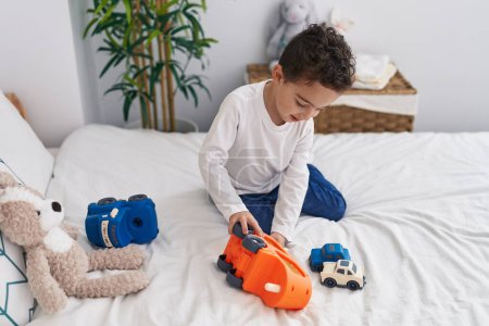 Foto de Adorable chico hispano jugando con juguete de coche sentado en la cama en el dormitorio - Imagen libre de derechos