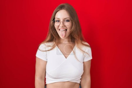Foto de Joven mujer caucásica de pie sobre fondo rojo sacando la lengua feliz con expresión divertida. concepto de emoción. - Imagen libre de derechos
