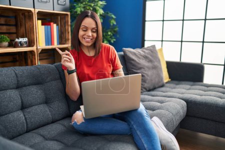 Photo for Young hispanic woman using laptop at home smiling happy pointing with hand and finger to the side - Royalty Free Image