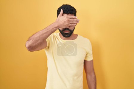 Téléchargez les photos : Homme hispanique avec la barbe debout sur fond jaune couvrant les yeux avec la main, l'air grave et triste. concept de non-visibilité, de dissimulation et de rejet - en image libre de droit