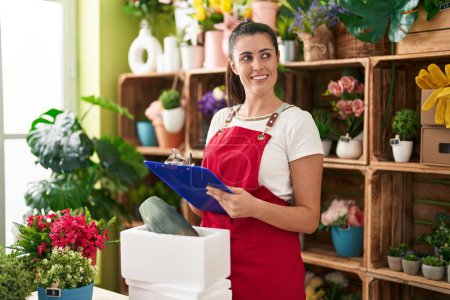 Photo for Young beautiful hispanic woman florist unpacking plant jar writing on document at flower shop - Royalty Free Image