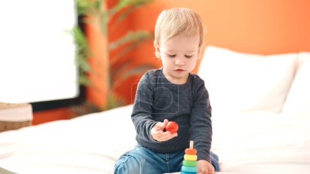 Téléchargez les photos : Adorable tout-petit blond jouant avec des cerceaux jouet assis sur le lit dans la chambre - en image libre de droit