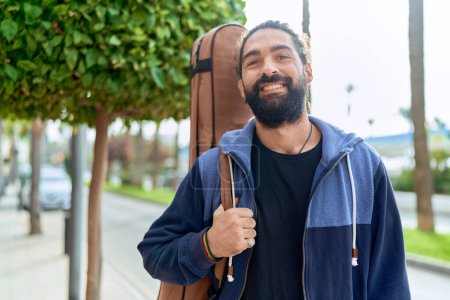Téléchargez les photos : Young hispanic man musician smiling confident holding guitar case at street - en image libre de droit