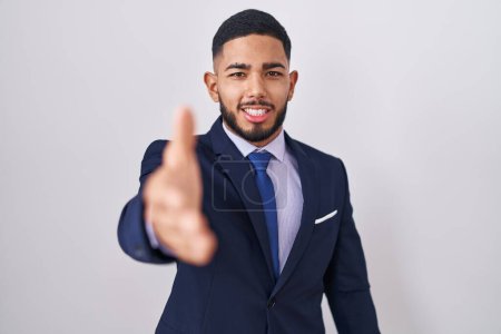 Foto de Joven hombre hispano con traje de negocios y corbata sonriente amable ofreciendo apretón de manos como saludo y bienvenida. negocios exitosos. - Imagen libre de derechos