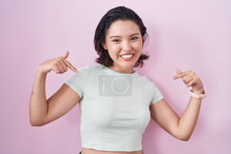 Foto de Mujer joven hispana de pie sobre fondo rosa mirando confiado con sonrisa en la cara, señalándose con los dedos orgullosos y felices. - Imagen libre de derechos