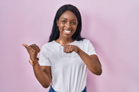 Foto de Mujer joven africana con camiseta blanca casual apuntando hacia atrás con la mano y los pulgares hacia arriba, sonriendo confiada - Imagen libre de derechos