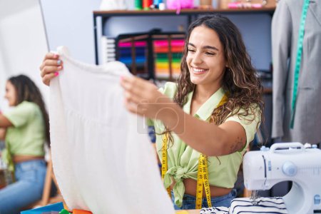 Foto de Joven hermosa mujer hispana sastre sonriendo confiado sosteniendo camiseta en la fábrica de ropa - Imagen libre de derechos
