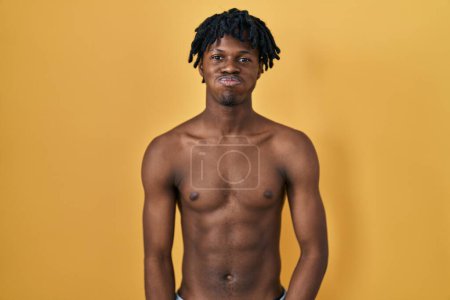 Foto de Hombre africano joven con rastas de pie sin camisa soplando mejillas con cara divertida. boca hinchada de aire, expresión loca. - Imagen libre de derechos
