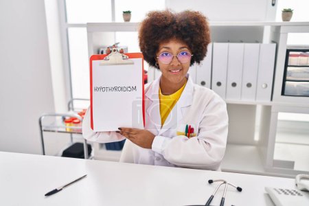 Photo for African american woman wearing doctor uniform holding clipboard with hypothyroidism message at clinic - Royalty Free Image