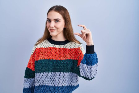 Photo for Young hispanic girl standing over blue background smiling and confident gesturing with hand doing small size sign with fingers looking and the camera. measure concept. - Royalty Free Image