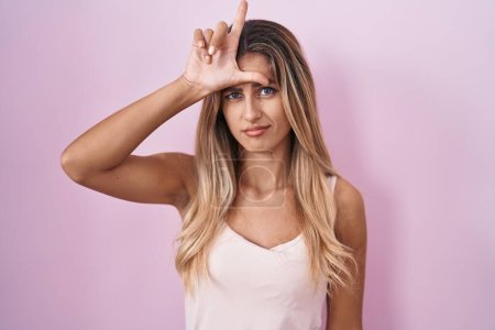 Téléchargez les photos : Young blonde woman standing over pink background making fun of people with fingers on forehead doing loser gesture mocking and insulting. - en image libre de droit