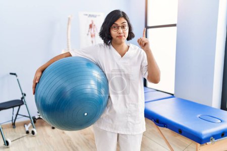 Photo for Young hispanic physiotherapist woman holding pilates ball smiling with an idea or question pointing finger with happy face, number one - Royalty Free Image