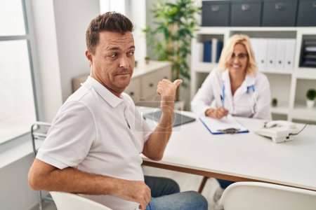 Photo for Hispanic man at the doctor pointing thumb up to the side smiling happy with open mouth - Royalty Free Image