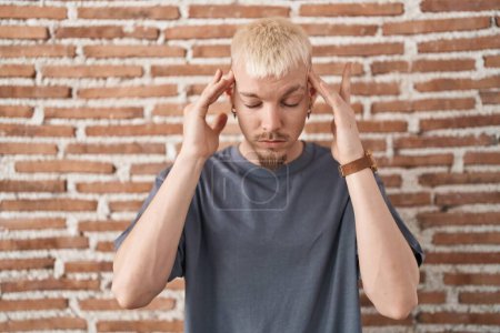 Téléchargez les photos : Jeune homme caucasien debout au-dessus du mur de briques avec la main sur la tête, maux de tête parce que le stress. migraine souffrant. - en image libre de droit