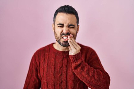 Téléchargez les photos : Jeune homme hispanique avec barbe portant un pull décontracté sur fond rose touchant la bouche avec la main avec une expression douloureuse à cause de maux de dents ou de maladies dentaires sur les dents. dentiste - en image libre de droit
